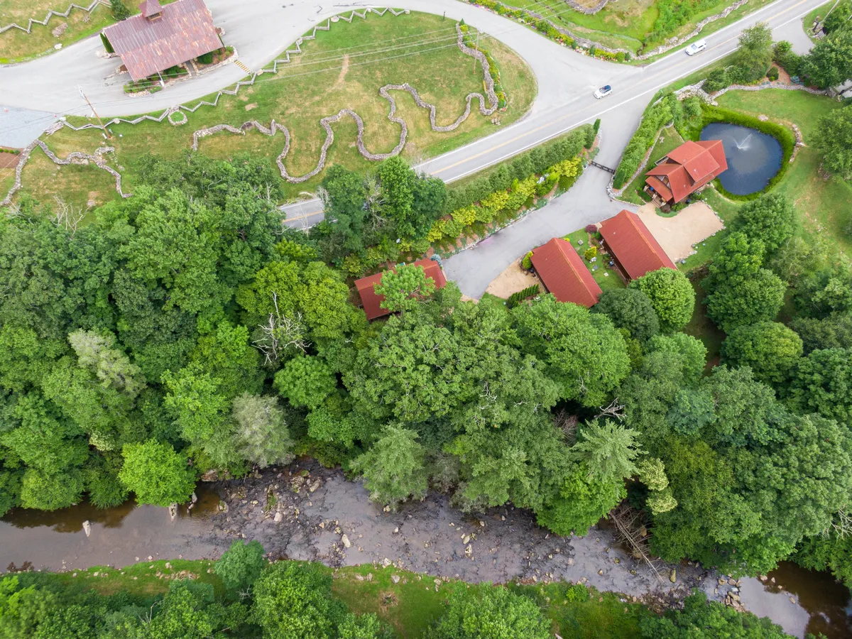 A picturesque scene featuring cabins with red roofs nestled beside a meandering stream, surrounded by lush greenery and a winding road. The setting suggests easy access to water and trails, inviting guests to explore the natural landscape.