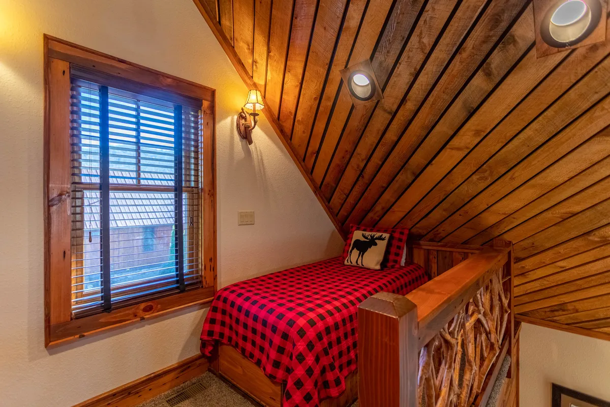 A warmly lit bedroom with a sloped wooden ceiling, a checkered bedspread, and a window with blinds offering a glimpse of the tranquil outdoors. The ambiance is intimate and welcoming, encouraging relaxation and quiet moments.