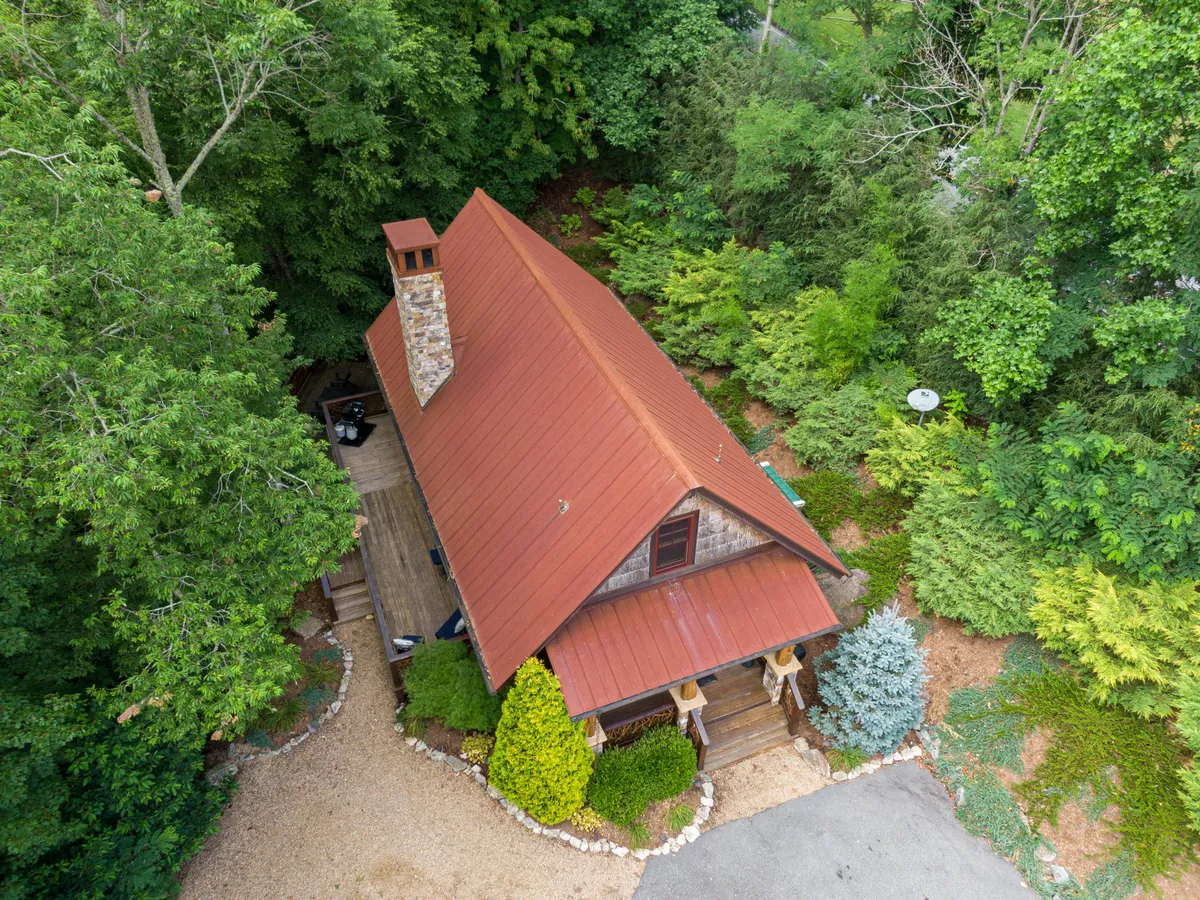 A wide view of the cabin's exterior, featuring a striking red metal roof, a stone chimney, and lush greenery enveloping the house. A gravel pathway leads towards the entrance, suggesting ease of access and a tranquil environment ideal for outdoor gatherings or setting off on local adventures.