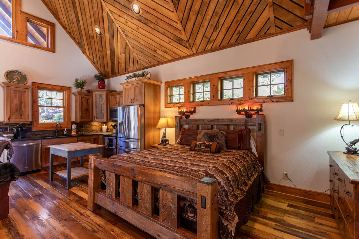 The image highlights a kitchen area with a visible wooden bed frame, sliding windows, ample lighting, and adjacent seating. The space is defined by its rustic appeal, with a warm color palette and homey touches conducive to cooking, socializing, and relaxing.