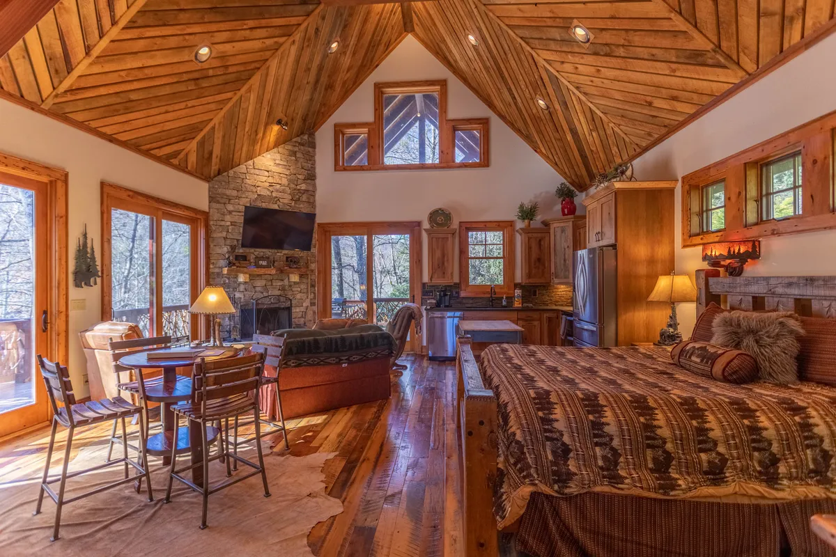 The image showcases a warm, inviting open-plan space featuring a vaulted wooden ceiling, a stacked stone fireplace, large windows that let natural light flood in, a dining table, and a bed with a patterned throw in the background. The rustic yet high-end furnishings and prominent wood accents evoke a lodge-inspired atmosphere, while the windows offer tranquil views of the outdoors, reinforcing the mountain retreat theme.