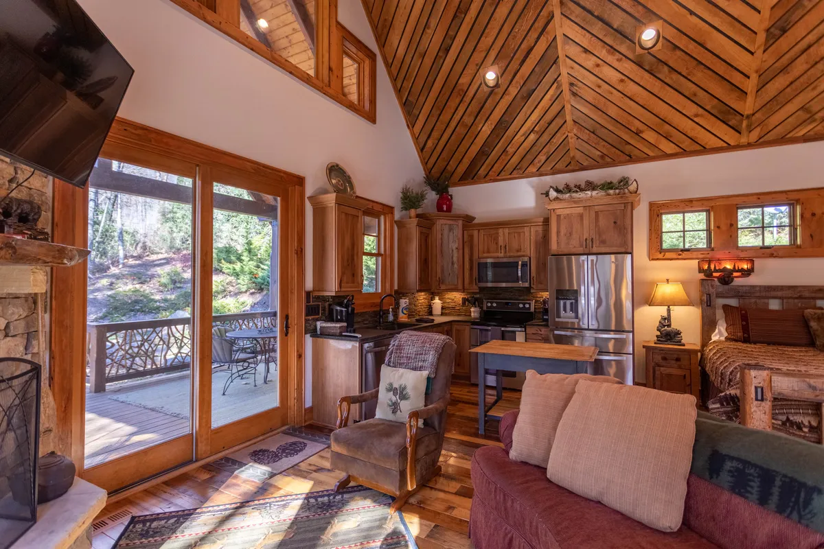 This image features a kitchen area with large windows offering an inviting outdoor view, rustic wooden kitchen cabinets, a comfortable seating area, and decor that blends wood and stone elements. The open sightlines from the kitchen to the living space emphasize connectivity, and the overall mood is warm and welcoming.