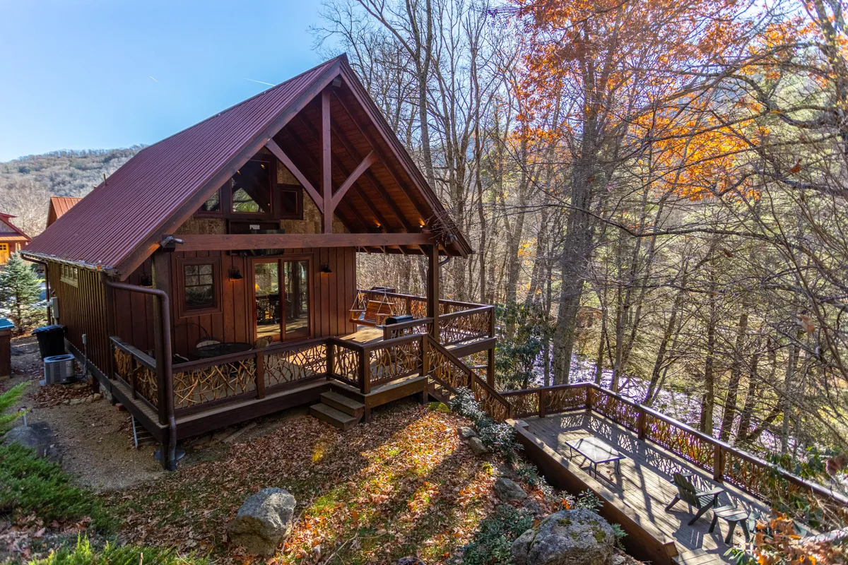 A charming wooden cabin with a deck furnished for seating, nestled among trees with mountain views in the background. The scene captures the possibility of relaxing on the deck, watching sunsets, or gathering around a fire pit.