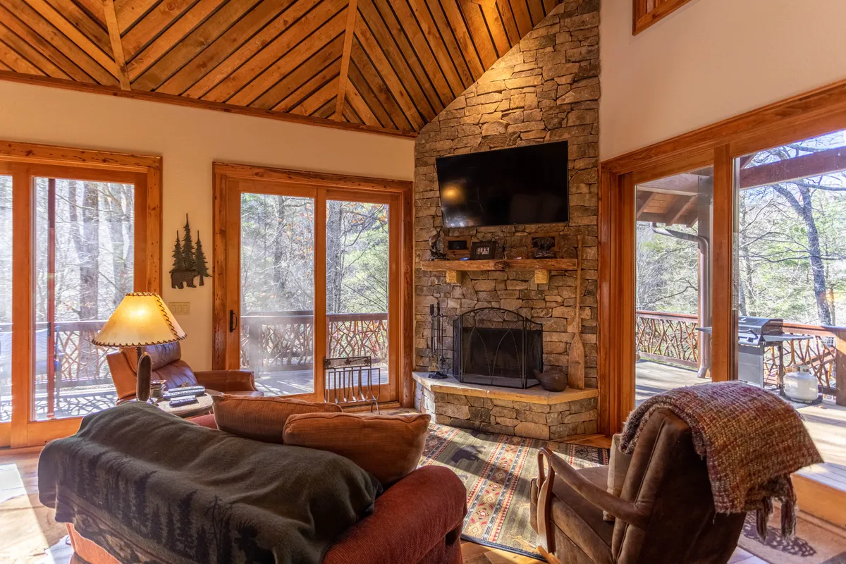 A cozy living room features a substantial stone fireplace as its focal point, flanked by large windows that draw in natural light and showcase the serene view outside. Comfortable seating is arranged around the fireplace, with visible wooden beams overhead enhancing the rustic, welcoming mood.