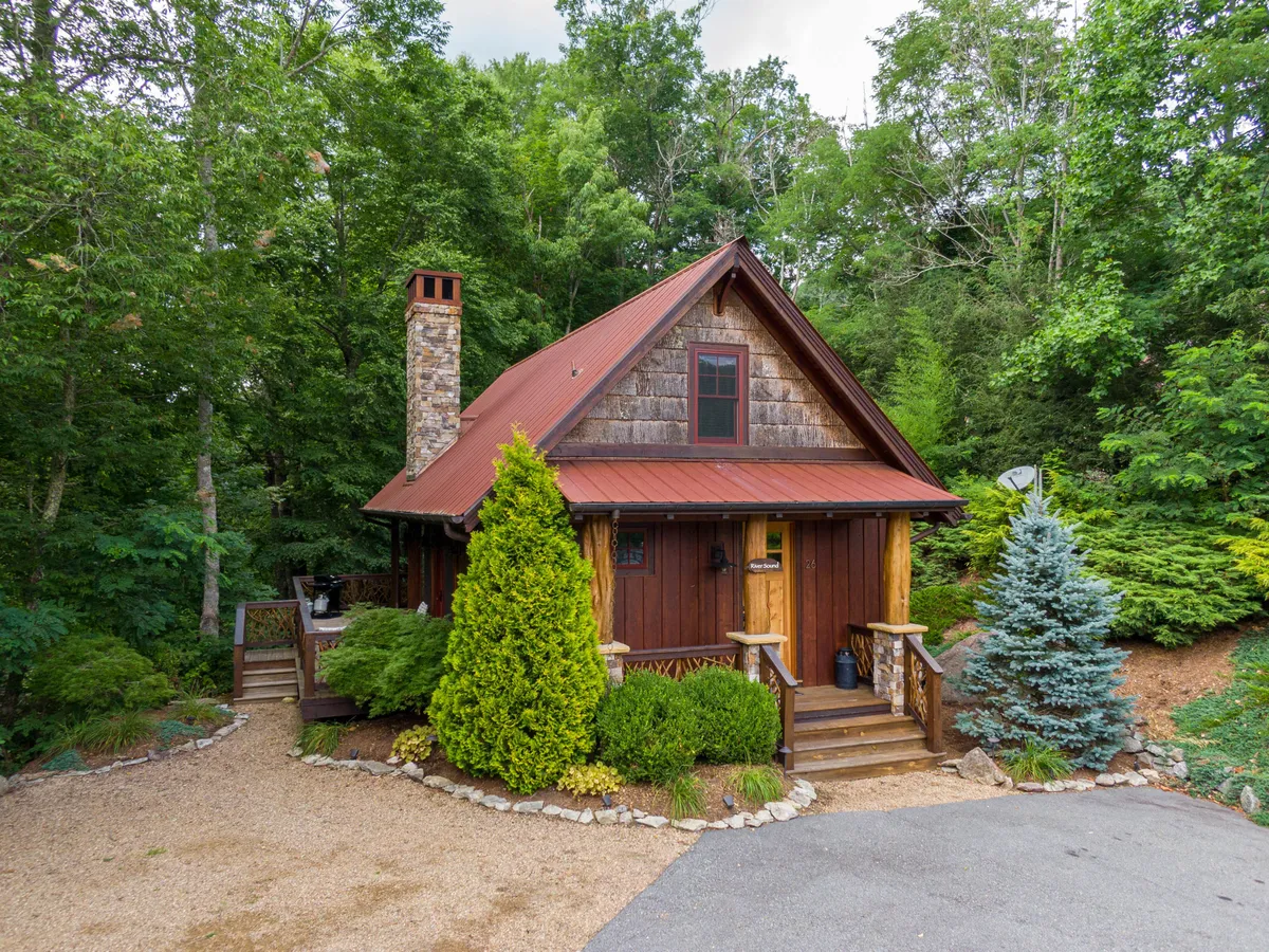 The image showcases a charming rustic wooden cabin nestled in lush greenery, with a prominent stone chimney and a welcoming, well-kept pathway leading up to the front porch. The inviting outdoor space hints at opportunities for stargazing, morning coffee, and enjoying the tranquil riverside setting, all surrounded by scenic Appalachian wilderness.