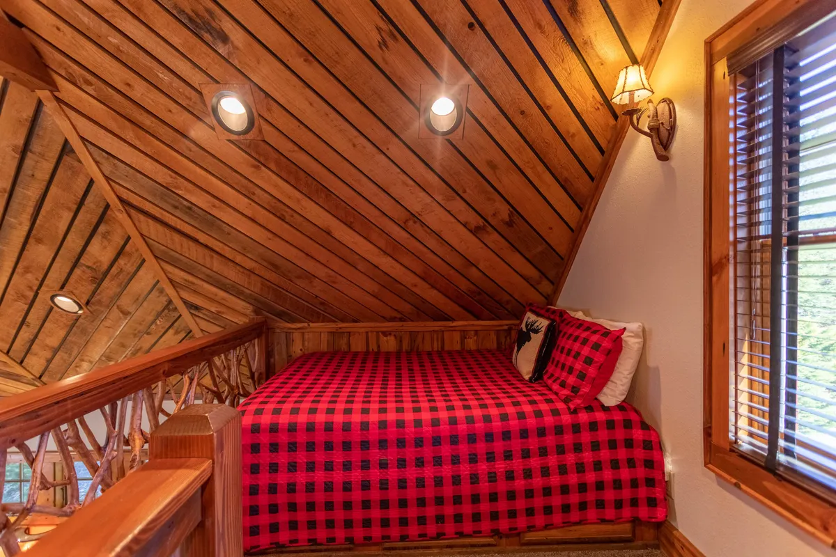 A cozy bedroom featuring plaid bedding, rustic wooden walls, and warm lighting from bedside lamps. The large window subtly suggests a view of the outdoors, reinforcing the connection to the natural surroundings and creating an inviting, restful retreat.
