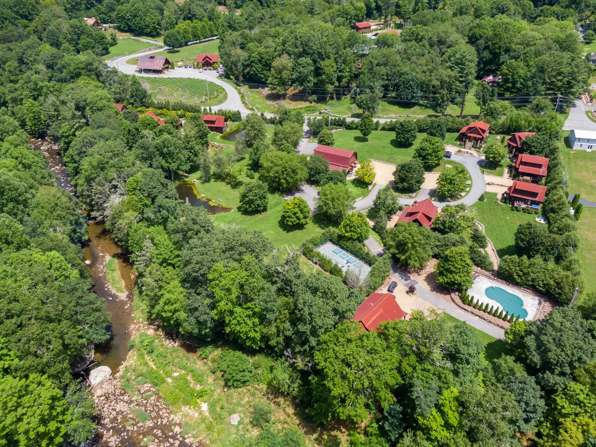 A wide-angle exterior view showing a winding river, several red-roofed cabins, a swimming pool, and dense greenery. The landscape is vibrant and teeming with natural features, indicating opportunities for swimming, hiking, fishing, and enjoying the changing seasons by the water.